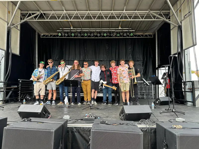 Members of The Scholars standing together on an outdoor stage with saxophones, trumpets, a bass guitar, and trombones