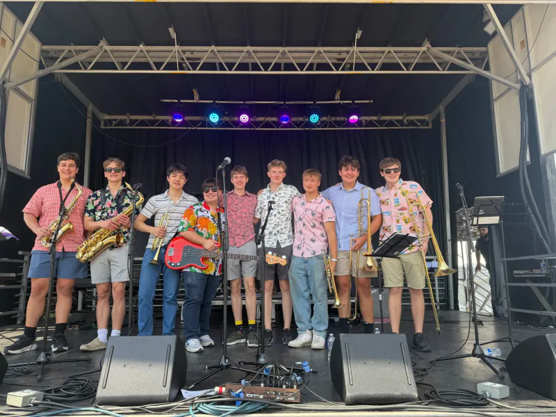 Members of The Scholars smiling on a festival stage with saxophones, trumpets, a bass guitar, and trombones
