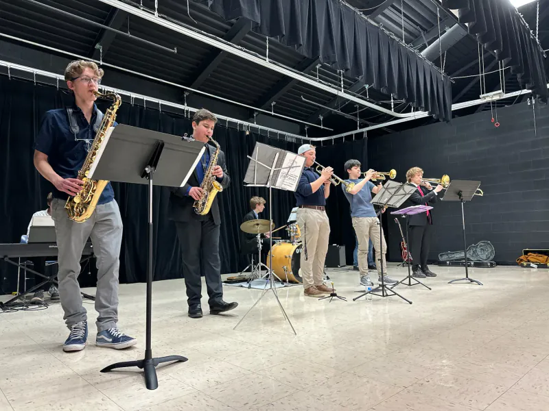 The Scholars playing in a room hung with black curtains: two saxophones in front, with keyboard, drums, and trumpets behind