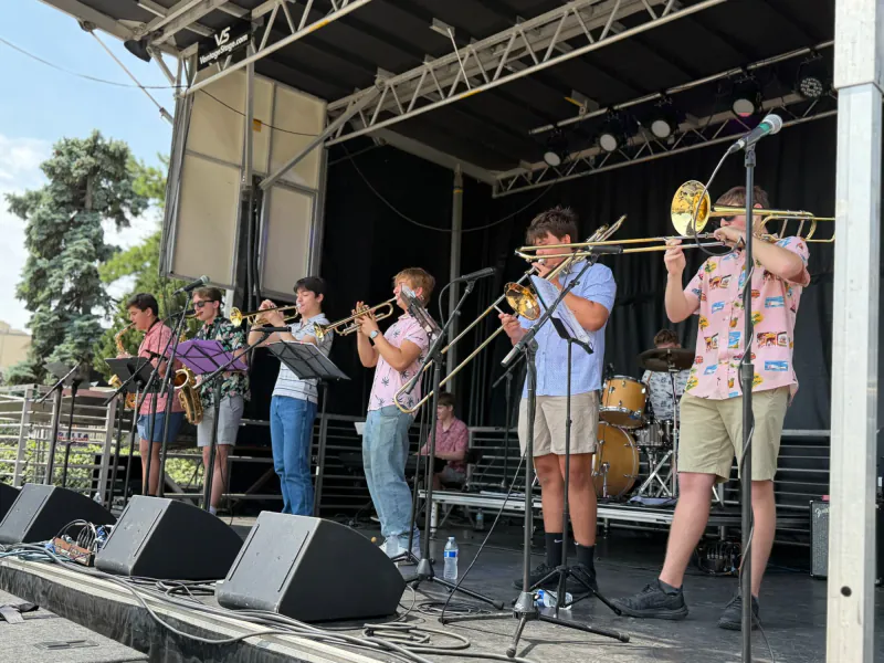 The Scholars playing on an outdoor festival stage: saxophones, trumpets, and trombones out front, with keyboard and drums behind