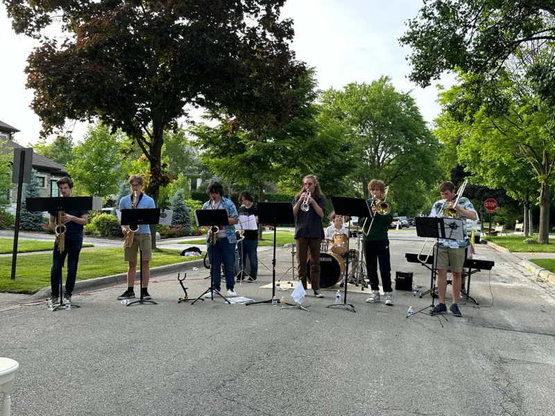The Scholars performing on a tree-lined residential street, with saxophones, trumpet, trombones, drums, and keyboard