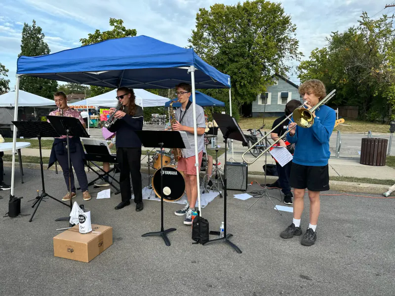 The Scholars playing under a blue canopy at an outdoor market, with a Scholars tip jar in front of the band